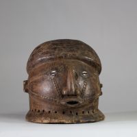 Masque-casque en bois, de forme hémisphérique Tabwa, RD congo. Visage sculpté avec un nez droit, saillant, de forme anguleuse, très caractéristique des styles Tabwa et Luba-Tabwa. Les yeux sont incrustés de cauris, Les scarifications faciales linéaires et parallèles, courant des tempes vers la bouche, sont typiques des traditions Tabwa. La patine naturelle, les abrasions autour de la base perforée