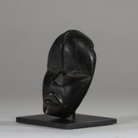 Masque passeport Dan, Côte d’Ivoire. Bois. Il présente une esthétique d’une grande pureté. Le visage, de forme ovale, est sculpté avec sobriété : les yeux sont mi-clos et incisés en amande, le nez est droit, triangulaire et structuré, et la bouche légèrement ouverte laisse deviner une expression intériorisée. Une ligne verticale en relief s’élève du haut du nez jusqu’au front, accentuant l’axe de