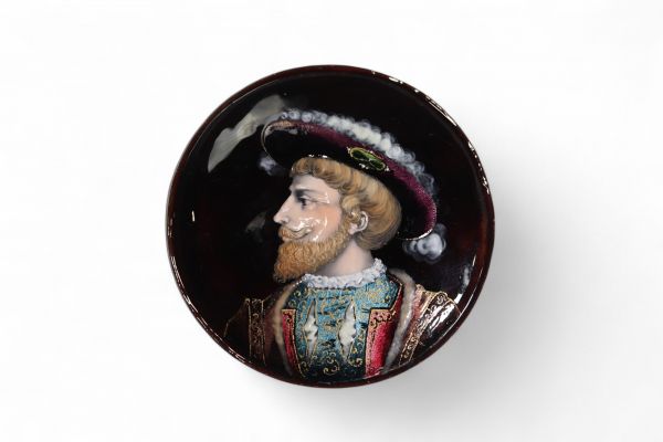 Médaillon en cuivre émaillé, Limoges, XIXᵉ siècle Médaillon circulaire en cuivre émaillé polychrome, représentant un portrait de Henri II, roi de France, de profil à gauche, vêtu d’un riche pourpoint bleu brodé or et coiffé d’un large chapeau à plume. Le fond grenat profond, l’élégance du visage et le raffinement des costumes évoquent les portraits de cour du XVIᵉ siècle. Le rendu minutieux de l’é