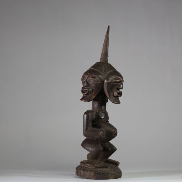 Puissant fétiche janiforme Songye, Congo RDC. Représentant une figure accroupie, aux bras anguleux et au torse tendu, coiffée d’une crête sagittale striée proéminente. La tête présente trois visages distincts disposés en triangle, chacun aux yeux mi-clos et à la bouche souriante, exprimant la vigilance et la vision multiple. Ce type de figure janiforme tripartite, rare dans l’art songye, renvoie à
