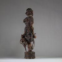 Intéressante statue féminine de type Bocio, Fon, Bénin. Le visage est sculpté de manière stylisée avec un nez proéminent, des yeux en amande profondément incisés, et des lèvres pleines. Une coiffe structurée orne la tête, enrichie de cauris incrustés et de perles. Le cou porte plusieurs rangs de colliers perlés. Le torse et le haut du corps sont entièrement recouverts de cauris patinés, suggérant