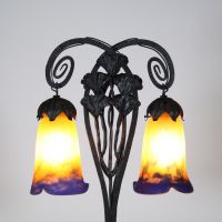 Muller Frères, Lunéville, vers 1905 Lampe à deux lumières en fer forgé martelé à décor de ginkgo biloba et verreries multicouches signées.  Superbe lampe Art Nouveau en fer forgé patiné noir, montée sur une base rectangulaire, s’élevant en double col de cygne à volutes stylisées. Le fût est orné de feuilles et hampes florales de ginkgo biloba, motif emblématique du symbolisme végétal de la Belle É