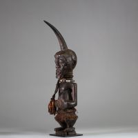 Fétiche Songye Nkisi, sous style Kalebwe, RD Congo. H : 46 cm. Figure masculine en bois, surmonté d’une corne rituelle, visage orné de clous de tapissier à fonction magique. Patine ancienne noire /brune, accessoires rituels suspendus (perles, fibres, cordelettes), cavités sous les bras. Nkisi de protection ou de justice activé par un « Nganga ». Style expressif et puissant propre aux Songye Kalebw
