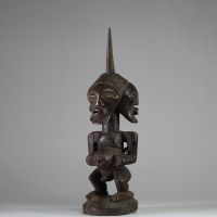 Puissant fétiche janiforme Songye, Congo RDC. Représentant une figure accroupie, aux bras anguleux et au torse tendu, coiffée d’une crête sagittale striée proéminente. La tête présente trois visages distincts disposés en triangle, chacun aux yeux mi-clos et à la bouche souriante, exprimant la vigilance et la vision multiple. Ce type de figure janiforme tripartite, rare dans l’art songye, renvoie à