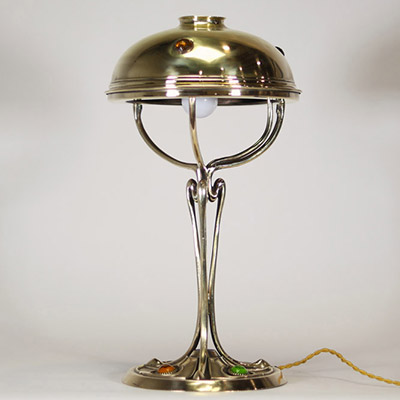 France, vers 1900 Lampe de table Art Nouveau en laiton, capeline ajourée et piétement à cabochons de verre coloré.  Cette lampe décorative de la période Art Nouveau présente une élégante silhouette élancée en laiton doré, avec un fût nervuré à lignes fluides convergeant vers une base circulaire décorée de cabochons de verre sertis en vert, rouge, ambre et jaune, typiques de l’ornementation polychr