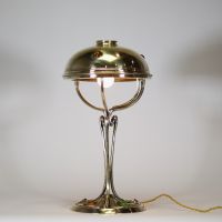 France, vers 1900 Lampe de table Art Nouveau en laiton, capeline ajourée et piétement à cabochons de verre coloré.  Cette lampe décorative de la période Art Nouveau présente une élégante silhouette élancée en laiton doré, avec un fût nervuré à lignes fluides convergeant vers une base circulaire décorée de cabochons de verre sertis en vert, rouge, ambre et jaune, typiques de l’ornementation polychr