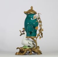 Fontaine à parfum, composée d’un vase en porcelaine de Chine émaillée bleu turquoise, datable du XVIIIe siècle, surmontée d’un couvercle orné d’une pomme de pin dorée. L’ensemble repose sur une base rocaille en bronze ciselé et doré, richement décorée de deux cygnes en porcelaine de Meissen, posés sur des tertres verts. Le décor est complété de fleurs en porcelaine polychrome, probablement de styl