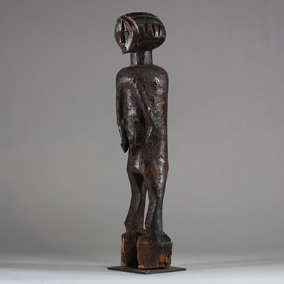 : Ancienne statue féminine Lwalwa, RD Congo. Bois, raphia. Le visage, de forme triangulaire inversée, nez rectiligne et saillant qui remonte jusqu’à la racine des cheveux. Yeux incisés,  bouche discrète et menton légèrement projeté. Le tronc, massif, est flanqué de deux bras longs et cylindriques, plaqués contre les flancs. Les seins sont petits et placés très bas, détail inhabituel qui renforce l
