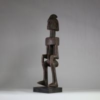 Statue masculine Dogon, Mali . Bois dense. Le style général est typique des productions Dogon anciennes, en particulier celles issues du plateau de Bandiagara. Le traitement géométrique du torse, l’élongation du cou, la posture figée et l’expression sobre indiquent une représentation d’ancêtre ou de figure spirituelle. La coiffe sagittale évoque les attributs cérémoniels du Hogon. La patine est an