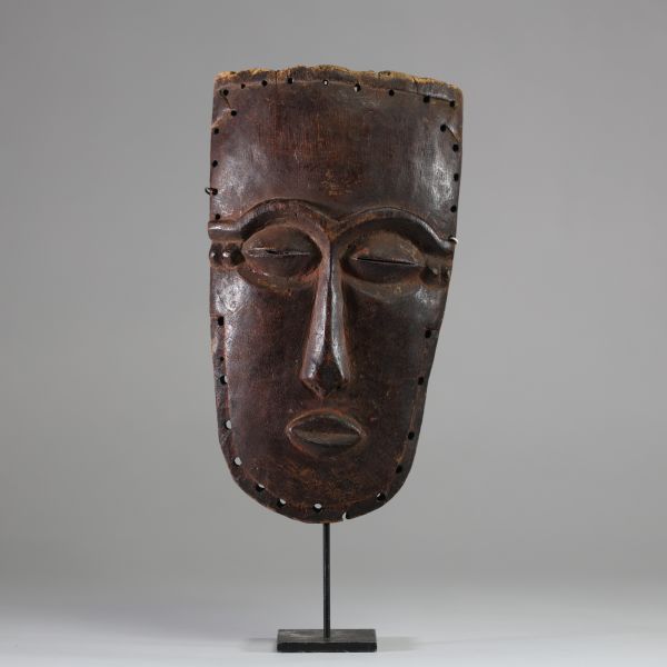 Masque facial anthropomorphe Lele ( bashilele), RD Congo, région du Kasaï. Bois présentant une surface patinée ancienne, plus brillante et lisse dans les zones exposées. Trous sur les bords permettant l’attache d’ornements ou a une structure. Yeux sculptés en amande avec fente étroite. La bouche est petite et sculptée en légère saillie. Le nez est droit et allongé avec une base marquée. Exemplaire
