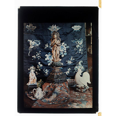 Léon Herlant (1873-1968) — 1 autochrome sur plaque Lumière, 12 x 9 cm.  Dans cette composition minimaliste d’une grande élégance, réalisée vers 1910, se déploie un subtil équilibre entre spiritualité, esthétique et culture de l’objet. Au centre trône un Amida Bouddha debout, sculpté en bois doré, figure emblématique du bouddhisme japonais de la période Edo, porteur de paix intérieure et de sagesse