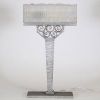 France, vers 1930 Lampe de bureau Art Déco en métal martelé nickelé et verre pressé à décor floral et géométrique.  Reposant sur une base rectangulaire, cette lampe présente un fût élancé à surface martelée, d’une sobriété élégante, souligné d’une finition nickelée mate. L’ensemble est rehaussé d’un nœud ornemental de volutes forgées à la main, typique du vocabulaire décoratif de l’époque.  L’abat
