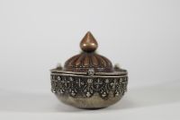 Kapala rituel, Tibet, XVIIIᵉ–XIXᵉ siècle Récipient rituel tibétain de type kapala, composé d’un corps en argent richement décoré de frises de têtes de kirtimukha, de chaînes perlées et de motifs stylisés. Le couvercle est en cuivre martelé, à décor gravé de pétales de lotus rayonnants, surmonté d’un prend finial en forme de flamme, symbole de sagesse dans le bouddhisme tantrique. Aux extrémités, d