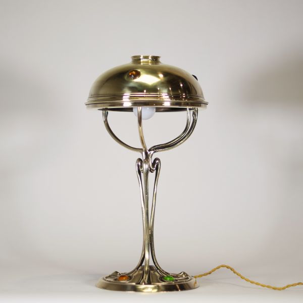 France, vers 1900 Lampe de table Art Nouveau en laiton, capeline ajourée et piétement à cabochons de verre coloré.  Cette lampe décorative de la période Art Nouveau présente une élégante silhouette élancée en laiton doré, avec un fût nervuré à lignes fluides convergeant vers une base circulaire décorée de cabochons de verre sertis en vert, rouge, ambre et jaune, typiques de l’ornementation polychr