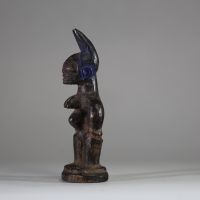 : Statuette Ibéji féminine, Yoruba, Nigéria. Figure féminine debout, dans une posture frontale rigide, les bras éloignés du corps reposant sur des hanches stylisées. Le visage est sculpté avec des yeux en amande, un front bombé, des scarifications linéaires, et une bouche aux lèvres striées horizontalement. Haute coiffe bifide . Patine noire ancienne avec des zones d’usure naturelle aux points de