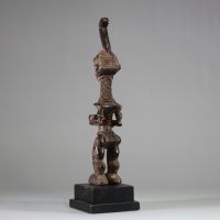 Statuette Bena Lulua, Région du Kasaï RD Congo. Bois. Très belle représentation d’ une figure féminine debout, aux proportions allongées. Tête légèrement surdimensionnée, ornée d'une coiffe cérémonielle monumentale se terminant en crosse verticale. Le visage montre des traits stylisés : yeux en amande, lèvres pleines, nez droit, et scarifications typiques sur le front, les joues et le menton. Le c
