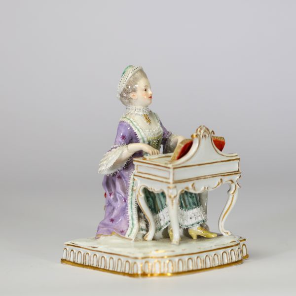 Figurine en porcelaine polychrome de la manufacture de Meissen « Jeune femme devant un petit piano ou clavecin. La base à décor moulé de lambrequins dorés repose sur un socle rectangulaire. Marque aux