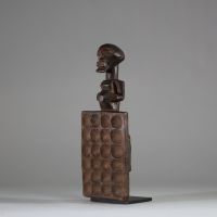 Plateau de jeu type « awalé » rituel Songye, RD Congo. Bois ancien, figure anthropomorphe stylisée, dont la partie inférieure est un plateau creusé de 24 alvéoles disposées en quatre rangées de six. Le personnage reprend les canons classiques de la culture Songye avec les bras collés au corps, et un ventre proéminent. Bel objet initiatique et symbolique à la croisée du ludique et du sacral, associ