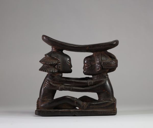 Ancien appui-nuque Luba « Shankadi », Congo RDC. Très bel exemplaire représentant deux figures humaines assises face à face, les bras posés sur les épaules de l’autre, dans une posture d’étreinte rituelle. Chacune soutient une tablette repose-nuque incurvée. L’une des figures présente une coiffure en croix (tulume) et l’autre une coiffure en cascade ( dans la tradition du maître de Kalundwe) . Les