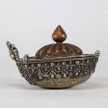 Kapala rituel, Tibet, XVIIIᵉ–XIXᵉ siècle Récipient rituel tibétain de type kapala, composé d’un corps en argent richement décoré de frises de têtes de kirtimukha, de chaînes perlées et de motifs stylisés. Le couvercle est en cuivre martelé, à décor gravé de pétales de lotus rayonnants, surmonté d’un prend finial en forme de flamme, symbole de sagesse dans le bouddhisme tantrique. Aux extrémités, d