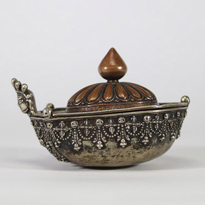Kapala rituel, Tibet, XVIIIᵉ–XIXᵉ siècle Récipient rituel tibétain de type kapala, composé d’un corps en argent richement décoré de frises de têtes de kirtimukha, de chaînes perlées et de motifs stylisés. Le couvercle est en cuivre martelé, à décor gravé de pétales de lotus rayonnants, surmonté d’un prend finial en forme de flamme, symbole de sagesse dans le bouddhisme tantrique. Aux extrémités, d