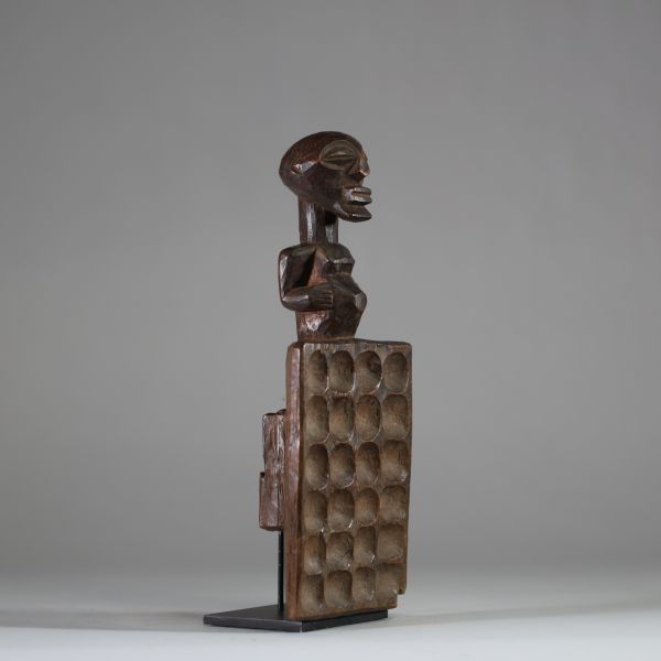 Plateau de jeu type « awalé » rituel Songye, RD Congo. Bois ancien, figure anthropomorphe stylisée, dont la partie inférieure est un plateau creusé de 24 alvéoles disposées en quatre rangées de six. Le personnage reprend les canons classiques de la culture Songye avec les bras collés au corps, et un ventre proéminent. Bel objet initiatique et symbolique à la croisée du ludique et du sacral, associ