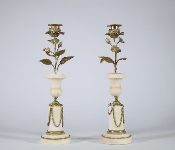 Paire de bougeoirs en marbre et bronze doré, travail de style Louis XVI Élégante paire de bougeoirs en marbre blanc veiné à base circulaire ornée de guirlandes en chaînette de bronze doré, la partie centrale prenant la forme d’un vase balustre, surmonté d’un décor végétal en bronze doré figurant une tige florale montée en candélabre, chaque fleur formant bobèche.  L’ensemble évoque avec fidélité l