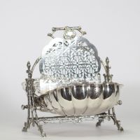 Élégante boîte à biscuits en métal argenté, à ouverture double, dite « shell biscuit box », prenant la forme d’un coquillage stylisé monté sur un piètement ajouré à motifs de branches naturalistes croisées. Le couvercle à charnières s’ouvre de part et d’autre pour dévoiler deux compartiments intérieurs à décor ajouré d’entrelacs floraux et géométriques. L’ensemble repose sur quatre pieds en forme