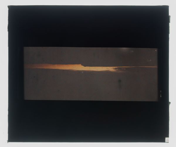 Léon Herlant (1873-1968) — 1 autochrome sur plaque Lumière, 8,5 x 10 cm.  Autochrome d'un coucher ou lever de soleil, circa 1910  Cet autochrome saisit un moment de transformation fugace, où la lumière et l'ombre se battent pour prendre forme. Le coucher ou lever du soleil est ici magnifiquement fragmenté par une falaise imposante et des nuages noirs qui coupent la scène en deux. L’horizon, partie