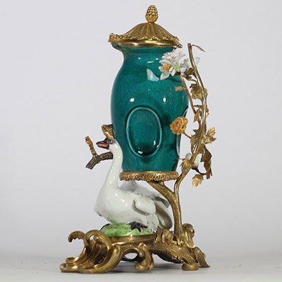 Fontaine à parfum, composée d’un vase en porcelaine de Chine émaillée bleu turquoise, datable du XVIIIe siècle, surmontée d’un couvercle orné d’une pomme de pin dorée. L’ensemble repose sur une base rocaille en bronze ciselé et doré, richement décorée de deux cygnes en porcelaine de Meissen, posés sur des tertres verts. Le décor est complété de fleurs en porcelaine polychrome, probablement de styl