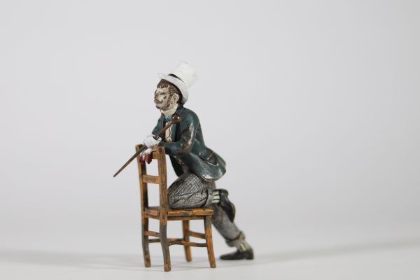 Bronze de Vienne, XIXᵉ siècle Petite figure en bronze polychrome représentant un homme assis à l’envers sur une chaise, vêtu d’un costume bourgeois avec chapeau haut-de-forme, gants blancs et canne. Il s’agit d’un exemple typique des bronzes de Vienne du XIXᵉ siècle, appréciés pour leurs sujets pittoresques, souvent inspirés du théâtre, des scènes de genre ou de la vie citadine.