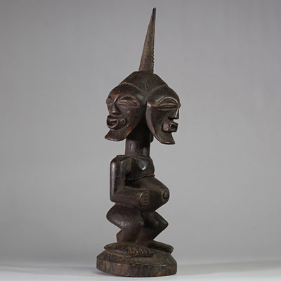 Puissant fétiche janiforme Songye, Congo RDC. Représentant une figure accroupie, aux bras anguleux et au torse tendu, coiffée d’une crête sagittale striée proéminente. La tête présente trois visages distincts disposés en triangle, chacun aux yeux mi-clos et à la bouche souriante, exprimant la vigilance et la vision multiple. Ce type de figure janiforme tripartite, rare dans l’art songye, renvoie à