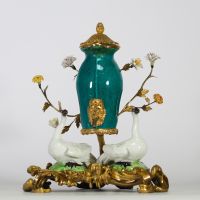 Fontaine à parfum, composée d’un vase en porcelaine de Chine émaillée bleu turquoise, datable du XVIIIe siècle, surmontée d’un couvercle orné d’une pomme de pin dorée. L’ensemble repose sur une base rocaille en bronze ciselé et doré, richement décorée de deux cygnes en porcelaine de Meissen, posés sur des tertres verts. Le décor est complété de fleurs en porcelaine polychrome, probablement de styl