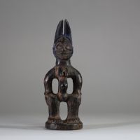: Statuette Ibéji féminine, Yoruba, Nigéria. Figure féminine debout, dans une posture frontale rigide, les bras éloignés du corps reposant sur des hanches stylisées. Le visage est sculpté avec des yeux en amande, un front bombé, des scarifications linéaires, et une bouche aux lèvres striées horizontalement. Haute coiffe bifide . Patine noire ancienne avec des zones d’usure naturelle aux points de
