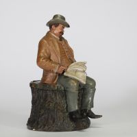 Bernard Bloch (atelier BB), Bohême, fin XIXe – début XXe siècle Pot à tabac en terre cuite polychrome figurant un homme assoupi vêtu d’un costume campagnard, assis sur une souche, les yeux mi-clos, un journal déployé sur les genoux.