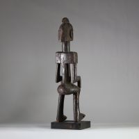 Statue masculine Dogon, Mali . Bois dense. Le style général est typique des productions Dogon anciennes, en particulier celles issues du plateau de Bandiagara. Le traitement géométrique du torse, l’élongation du cou, la posture figée et l’expression sobre indiquent une représentation d’ancêtre ou de figure spirituelle. La coiffe sagittale évoque les attributs cérémoniels du Hogon. La patine est an