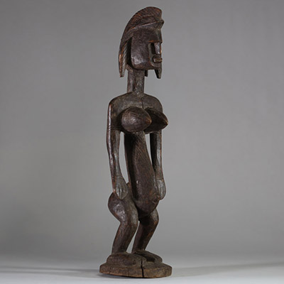 Statue rituelle Bamana « Jonyeleni » , Mali. Sculptée dans un bois ancien à patine brune, représentant une figure verticale aux traits stylisés : coiffe sagittale striée, nez droit, seins coniques scarifiés, ombilic et sexe proéminents. Les mains sont tendues le long du corps avec les paumes ouvertes, dans une posture typique des figures d’enseignement initiatique du Jon.La sculpture présente une