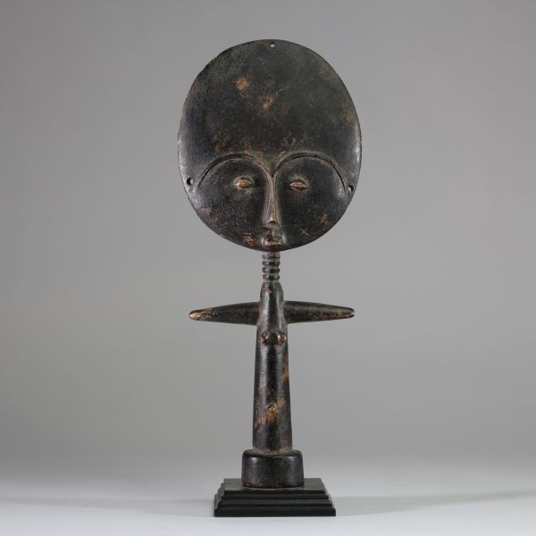 Poupée de fertilité Akua ba, groupe Akan, ethnie Ashanti, Ghana. Tête de forme plate et circulaire, très stylisée, yeux en amande, le nez fin et une petite bouche schématique. Colonne cylindrique avec les bras en croix et base évasée pour la stabilité. Patine riche et matte avec des variations subtiles de couleur, ce qui laisse présager une utilisation sur une longue période. L'arrière du disque d
