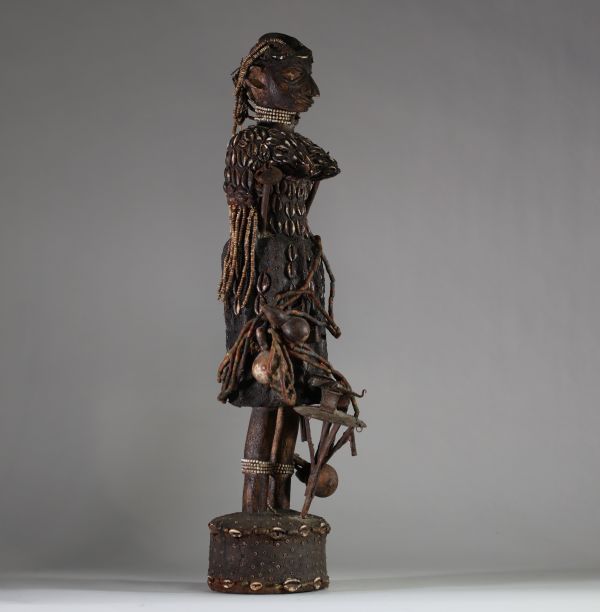 Intéressante statue féminine de type Bocio, Fon, Bénin. Le visage est sculpté de manière stylisée avec un nez proéminent, des yeux en amande profondément incisés, et des lèvres pleines. Une coiffe structurée orne la tête, enrichie de cauris incrustés et de perles. Le cou porte plusieurs rangs de colliers perlés. Le torse et le haut du corps sont entièrement recouverts de cauris patinés, suggérant