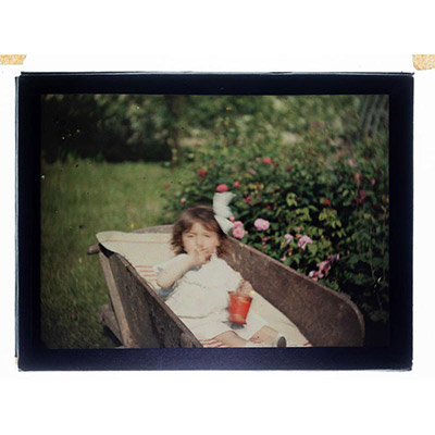 Léon Herlant (1873-1968) — autochrome sur plaque Lumière», 12 x 9 cm.  Petite fille en mouvement dans une brouette — Jardin fleuri Dans un jardin éclatant de fleurs roses, une petite fille vêtue de blanc apparaît en léger flou, saisie dans le mouvement vif d’une promenade en brouette. Une touche de rouge, discrète mais vibrante, vient accentuer la scène, apportant un contraste subtil à l’harmonie