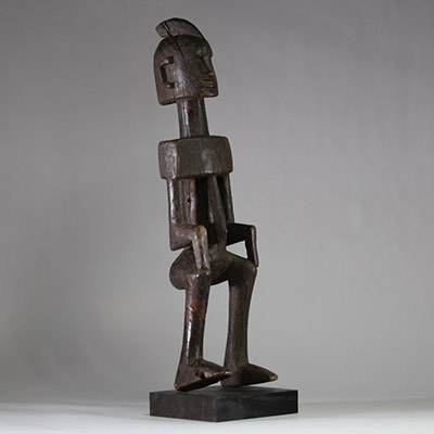 Statue masculine Dogon, Mali . Bois dense. Le style général est typique des productions Dogon anciennes, en particulier celles issues du plateau de Bandiagara. Le traitement géométrique du torse, l’élongation du cou, la posture figée et l’expression sobre indiquent une représentation d’ancêtre ou de figure spirituelle. La coiffe sagittale évoque les attributs cérémoniels du Hogon. La patine est an