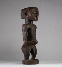 Statue Hemba Singiti, RD Congo. La statue représente une figure masculine debout, dans une posture frontale hiératique. La tête, surdimensionnée, est marquée par une barbe trapézoïdale finement striée, un visage calme aux yeux mi-clos, un nez droit et une bouche discrète. À l’arrière du crâne, une coiffe cruciforme profondément sculptée suggère une coiffure cérémonielle typique. Les bras sont plié