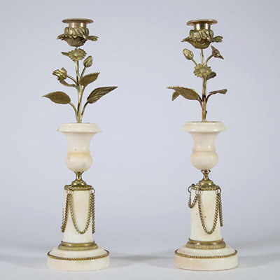 Paire de bougeoirs en marbre et bronze doré, travail de style Louis XVI Élégante paire de bougeoirs en marbre blanc veiné à base circulaire ornée de guirlandes en chaînette de bronze doré, la partie centrale prenant la forme d’un vase balustre, surmonté d’un décor végétal en bronze doré figurant une tige florale montée en candélabre, chaque fleur formant bobèche.  L’ensemble évoque avec fidélité l