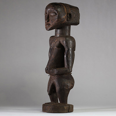 Statue Hemba Singiti, RD Congo. La statue représente une figure masculine debout, dans une posture frontale hiératique. La tête, surdimensionnée, est marquée par une barbe trapézoïdale finement striée, un visage calme aux yeux mi-clos, un nez droit et une bouche discrète. À l’arrière du crâne, une coiffe cruciforme profondément sculptée suggère une coiffure cérémonielle typique. Les bras sont plié