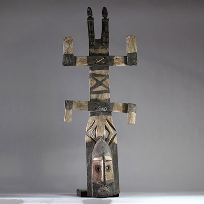 Grand masque Dogon « Kanaga », Mali. Bois ancien et sec, avec des pigments naturels noirs et blancs appliqués de manière traditionnelle. Le visage stylisé présente des yeux en triangle évidés, surmonté d'une superstructure composée de deux barres transversales et de deux personnages anthropomorphes sculptés au sommet. La structure générale respecte les canons de la statuaire Dogon liée aux cérémon