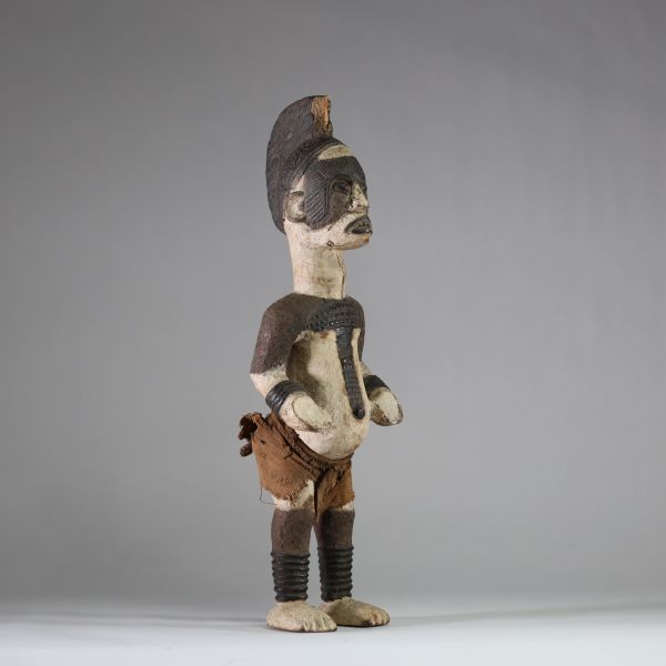 Statue Igbo « Alusi ». Nigéria. Bois, textile. Représentation masculine de type Alusi, divinité secondaire ou esprit tutélaire propre au peuple Igbo (sud-est du Nigeria), vénérée dans des sanctuaires collectifs ou familiaux. Grande crête sagittale sculptée d’une coiffe en éventail semi circulaire, ornée de motifs en spirales. Front bombé, yeux globuleux en amande, sans pupille, sourcils et joues m
