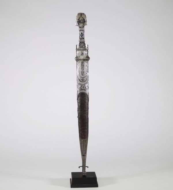 Poignard persan à décor d'argent, début XIXᵉ siècle Poignard oriental monté en argent repoussé et ciselé, présentant une garde et un fourreau richement ornés de motifs floraux et de cartouches géométriques, caractéristiques de l’art décoratif perse. Le pommeau bombé et la garde sont rehaussés de filets floraux, certains émaillés de bleu.  La lame à double tranchant, probablement forgée en acier, e