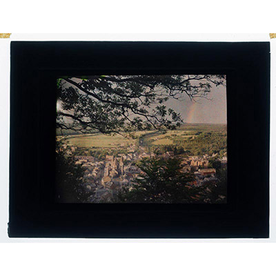 Léon Herlant (1873-1968) — 1 autochrome sur plaque Lumière, 8,5 x 10 cm.  Rare Autochrome d'un Arc-en-Ciel avec Vue sur la Ville de Spa, circa 1910  Cette rareté photographique est un autochrome exceptionnel, capturant un arc-en-ciel en plein éclat, avec la ville de Spa en arrière-plan, vue en plongée. Un instant naturel empreint de magie et de beauté fugace, sa richesse colorée est amplifiée par