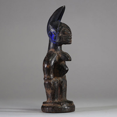 : Statuette Ibéji féminine, Yoruba, Nigéria. Figure féminine debout, dans une posture frontale rigide, les bras éloignés du corps reposant sur des hanches stylisées. Le visage est sculpté avec des yeux en amande, un front bombé, des scarifications linéaires, et une bouche aux lèvres striées horizontalement. Haute coiffe bifide . Patine noire ancienne avec des zones d’usure naturelle aux points de
