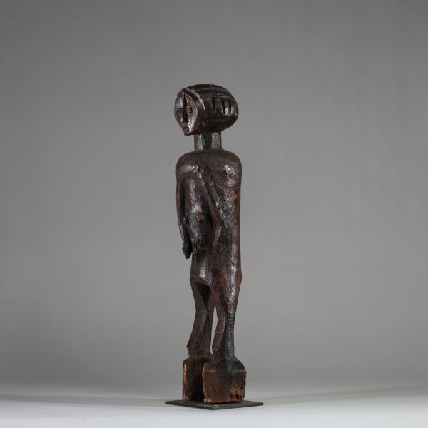 : Ancienne statue féminine Lwalwa, RD Congo. Bois, raphia. Le visage, de forme triangulaire inversée, nez rectiligne et saillant qui remonte jusqu’à la racine des cheveux. Yeux incisés,  bouche discrète et menton légèrement projeté. Le tronc, massif, est flanqué de deux bras longs et cylindriques, plaqués contre les flancs. Les seins sont petits et placés très bas, détail inhabituel qui renforce l
